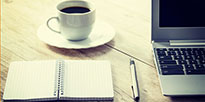 Symbolbild, auf dem eine Kaffeetasse, ein Notizblock mit Stift und ein Laptop zu sehen sind.